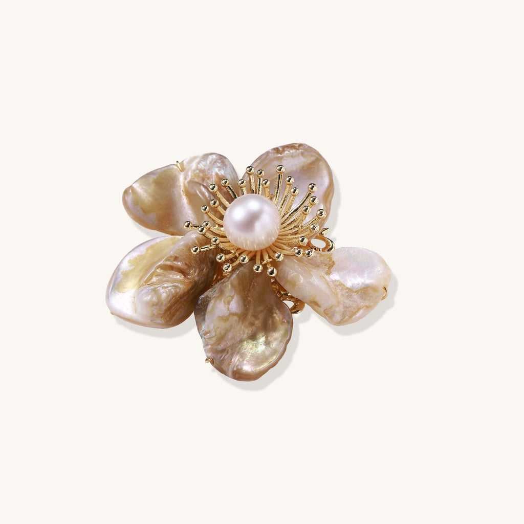 Elegant floral pin with peachy shell petals, gold radiating spokes, and white freshwater pearl center on cream background.