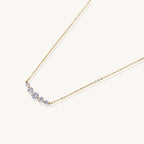 Delicate 14K gold plated necklace with curved arc of sparkling zirconia stones on cream background.